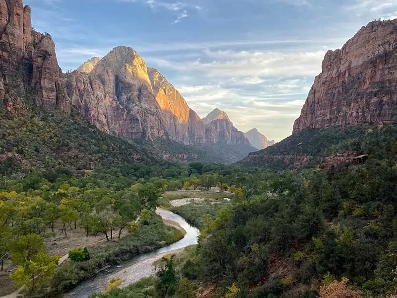 Scenic view of Zion National Park with a winding river, lush green trees, and towering rocky cliffs bathed in warm sunlight under a partly cloudy sky.